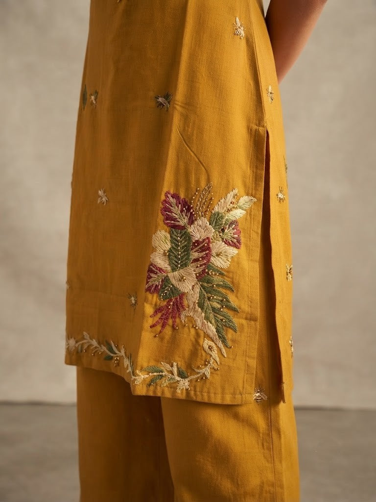 A woman modeling a mustard yellow kurta set with floral embroidery and tassel details, paired with matching wide-leg pants, against a neutral beige backdrop.