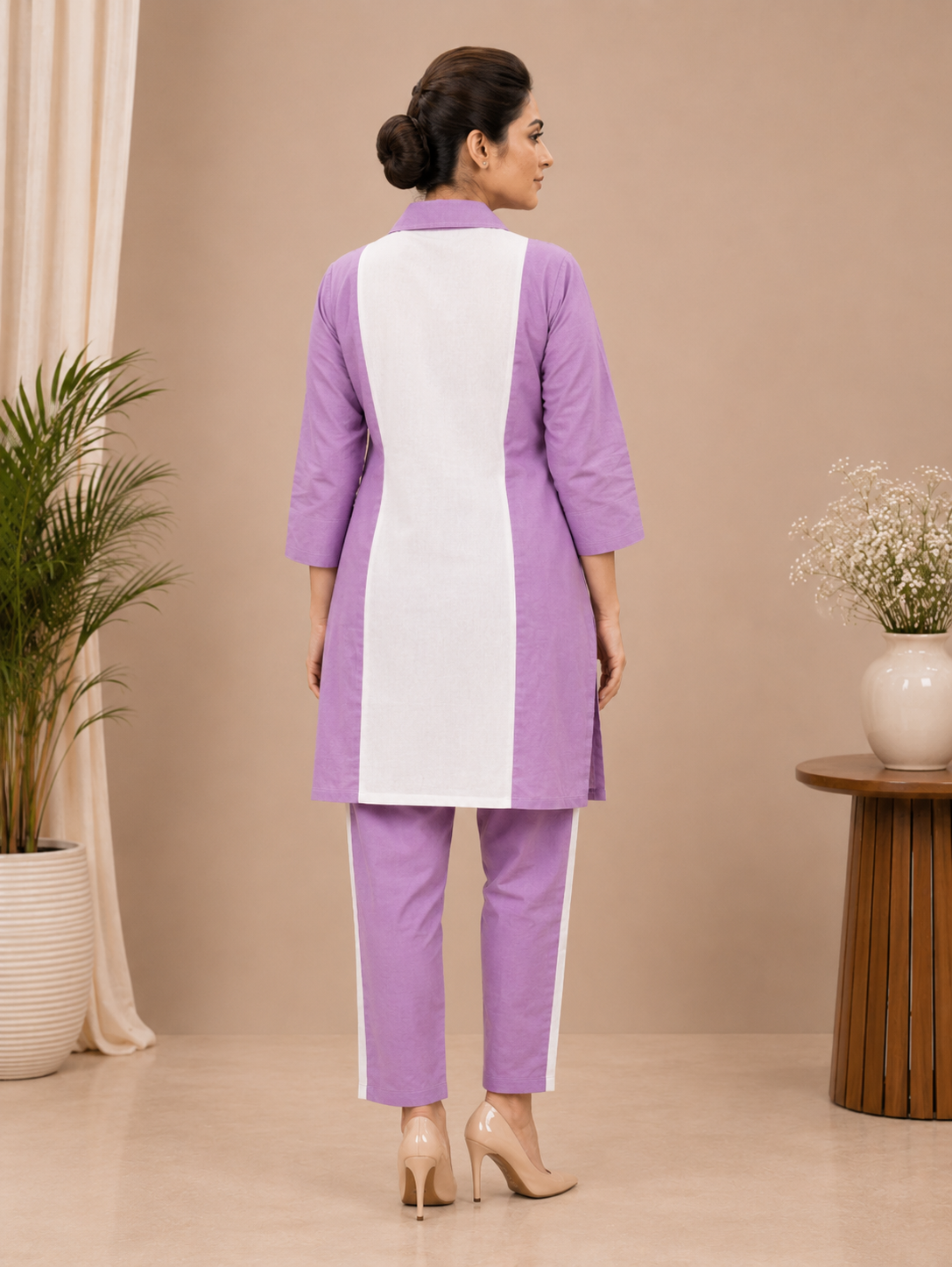 A woman modeling a lavender and white two-piece kurta set with purple floral embroidery, standing in a studio with beige walls and decorative plants.