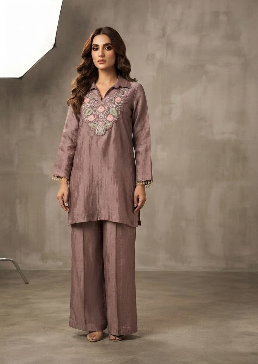 A woman modeling a dusty mauve two-piece kurta and trouser set with floral embroidery on the chest, standing against a textured gray studio backdrop.