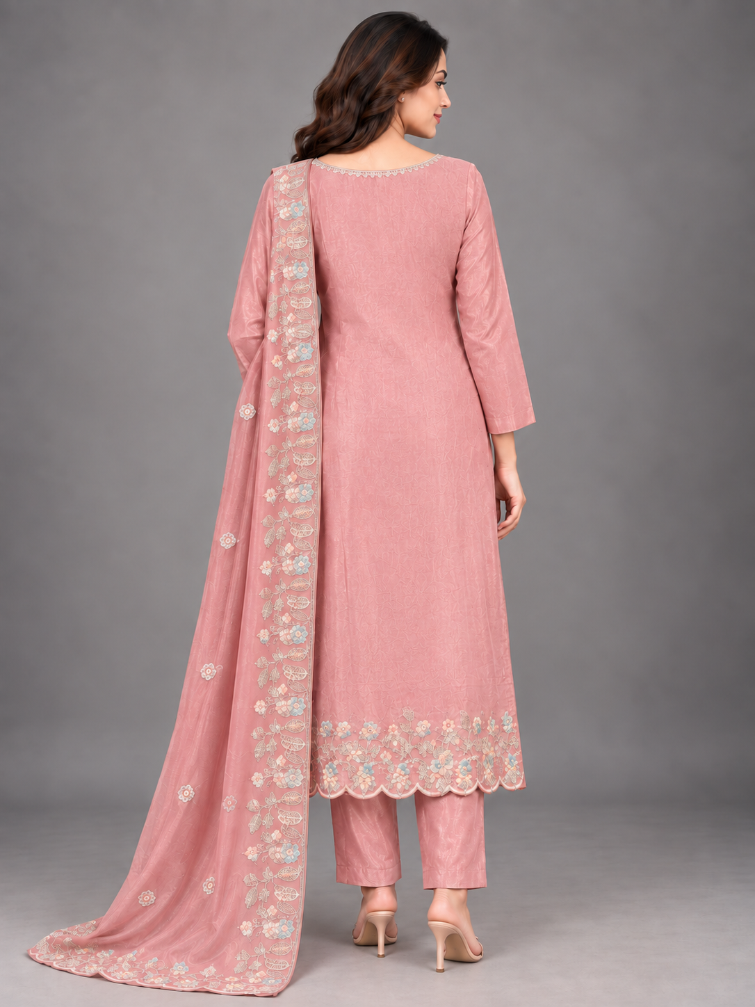 A woman modeling a pink traditional outfit with matching pants and a long sheer dupatta, featuring light blue and gold floral embroidery, against a solid gray studio background.