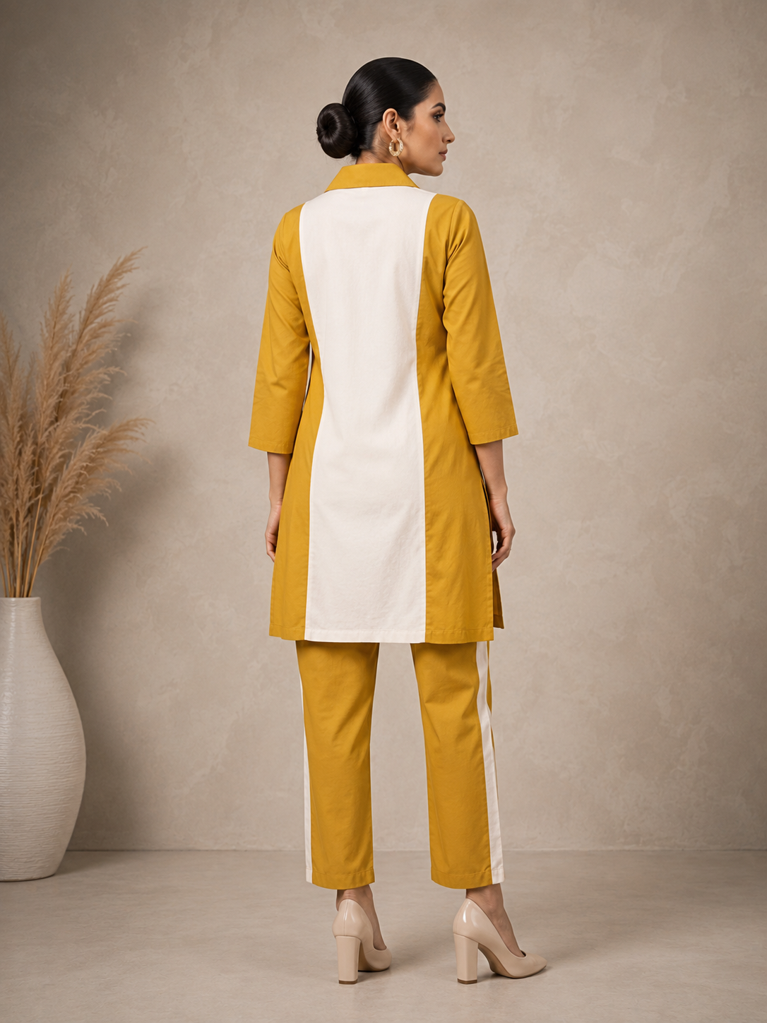 A woman models a mustard yellow and white kurta set with matching trousers, featuring yellow embroidered rose motifs on the white front panel. She is standing in a studio setting with a textured beige wall and a decorative vase of pampas grass on the left.