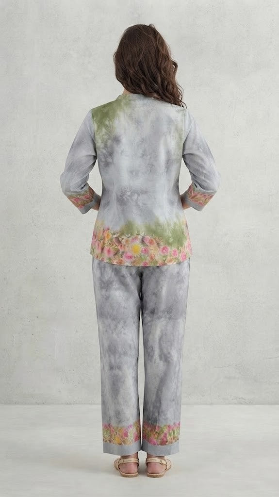 A woman modeling a two-piece traditional outfit featuring a light gray and white tie-dye kurta with green embroidery and colorful floral prints, paired with matching wide-leg trousers.