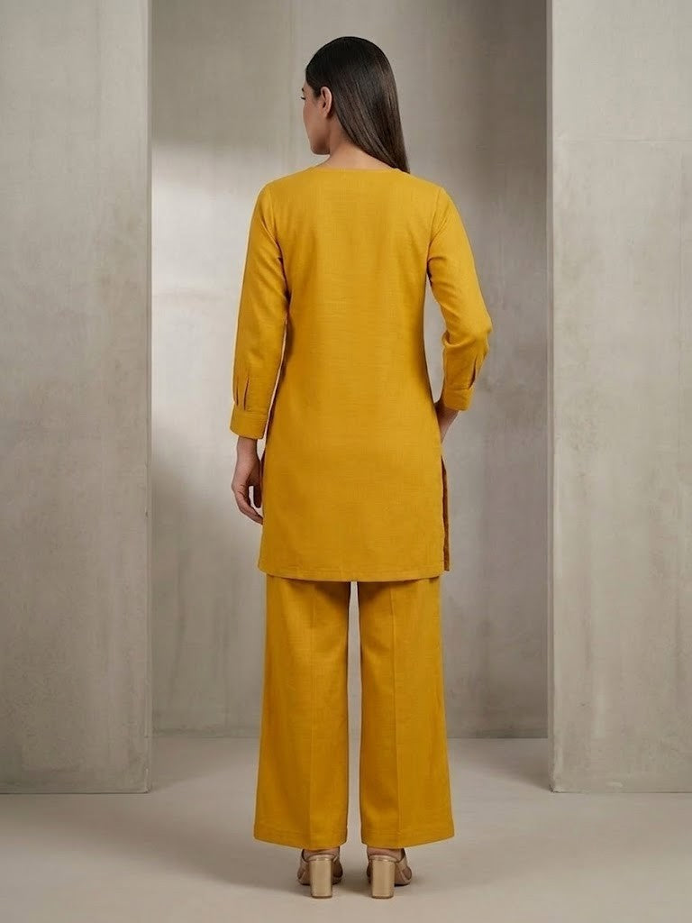 A woman modeling a mustard yellow two-piece kurta and trouser set with colorful floral embroidery on the sleeves and lower pant leg, standing in a minimalist concrete-walled studio.
