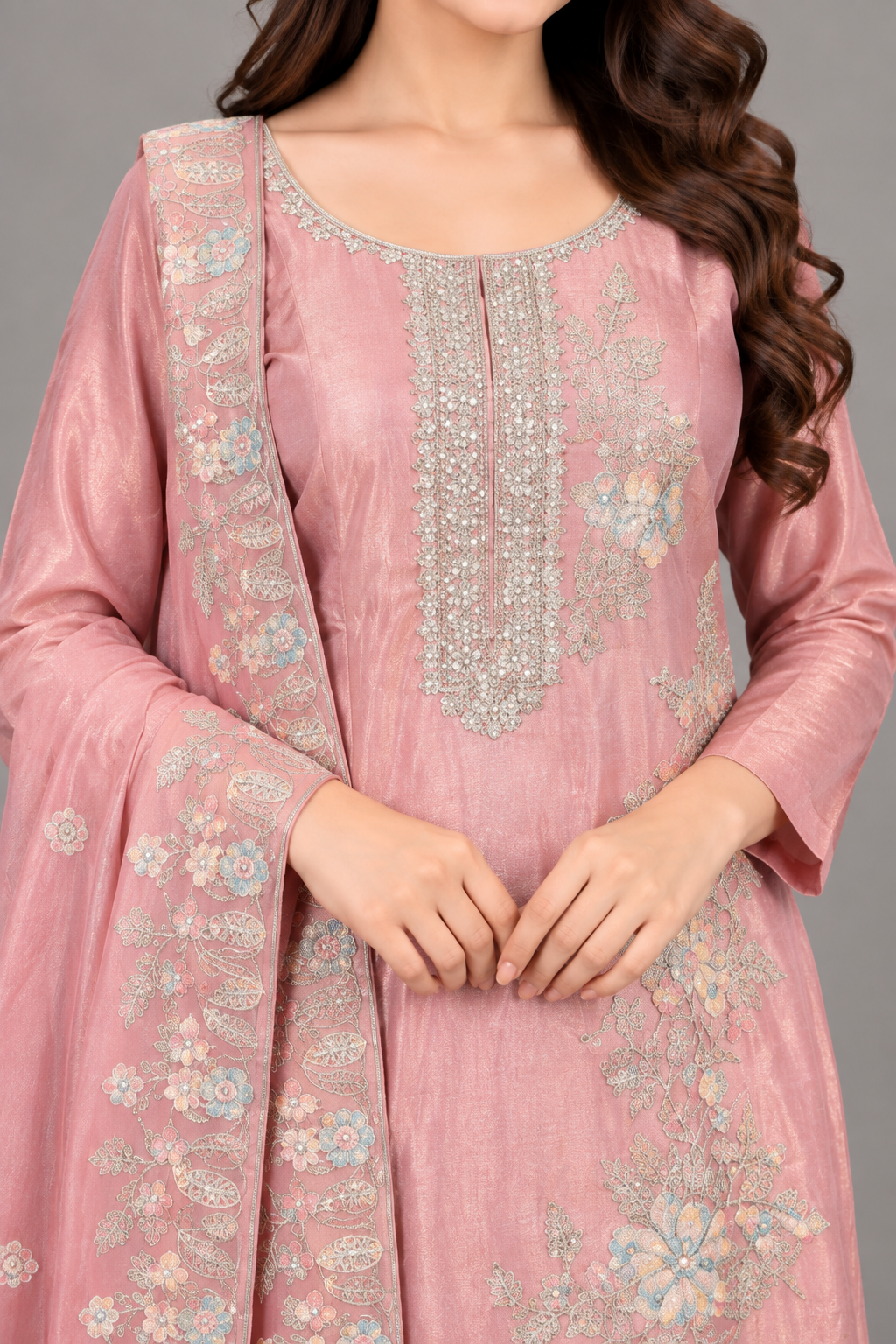 A woman modeling a pink traditional outfit with matching pants and a long sheer dupatta, featuring light blue and gold floral embroidery, against a solid gray studio background.