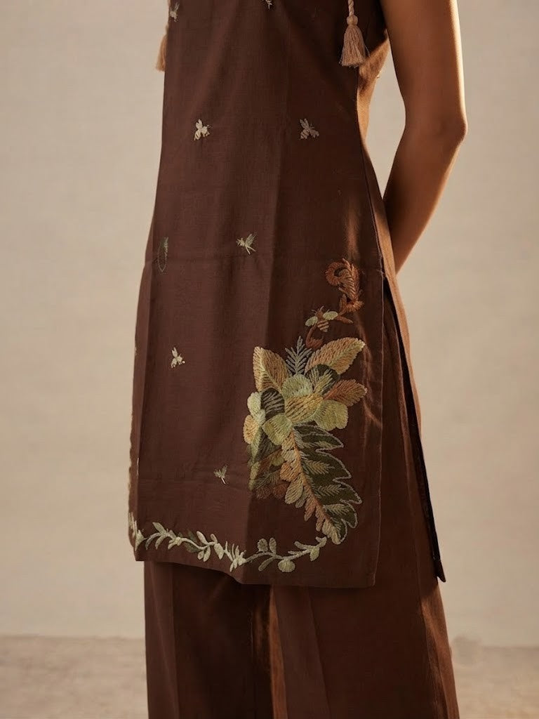 A woman modeling a dark brown sleeveless kurta with floral and butterfly embroidery, paired with matching wide-leg trousers, in a studio setting.
