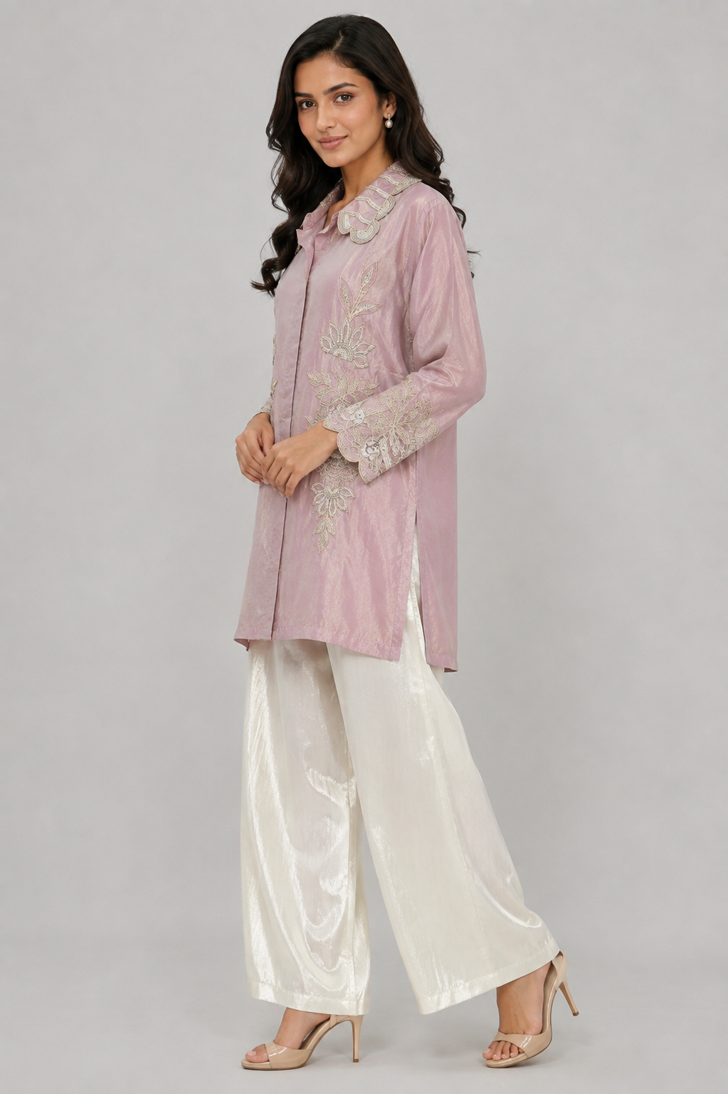 A woman modeling a light pink embroidered kurta paired with white satin palazzo pants and nude heels, set against a solid gray studio background.