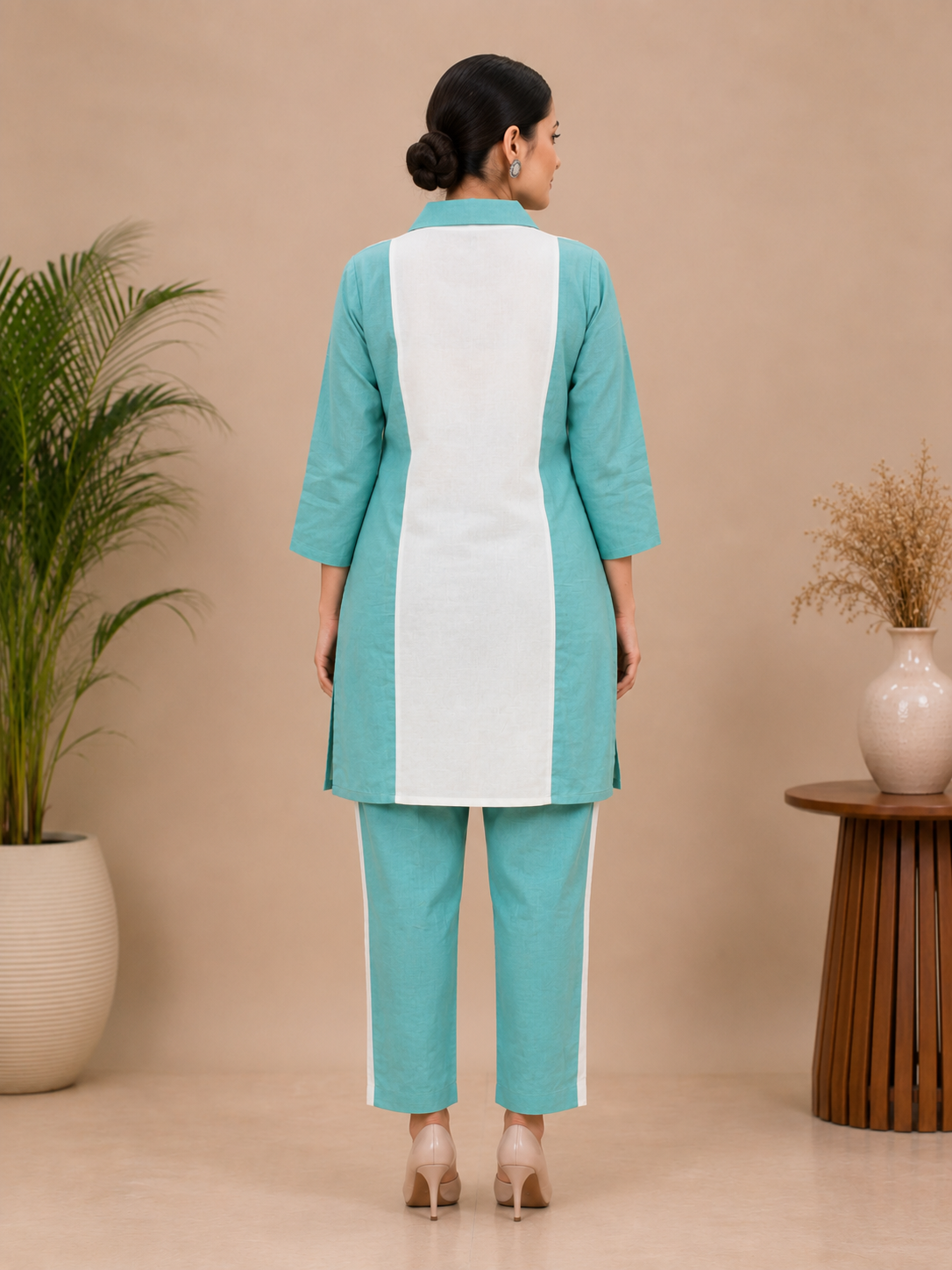 A woman modeling a two-piece kurta and trouser set in mint green and white, featuring a vertical white stripe on the kurta with two green floral prints, set against a warm beige background with decorative plants.