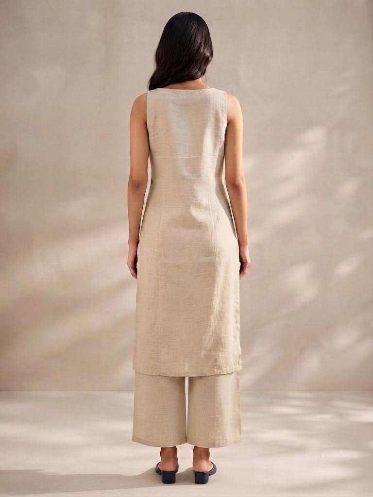 A woman modeling a sleeveless beige kurta with blue and green floral embroidery, paired with matching beige wide-leg pants and navy flats, set against a warm neutral background with soft shadows.