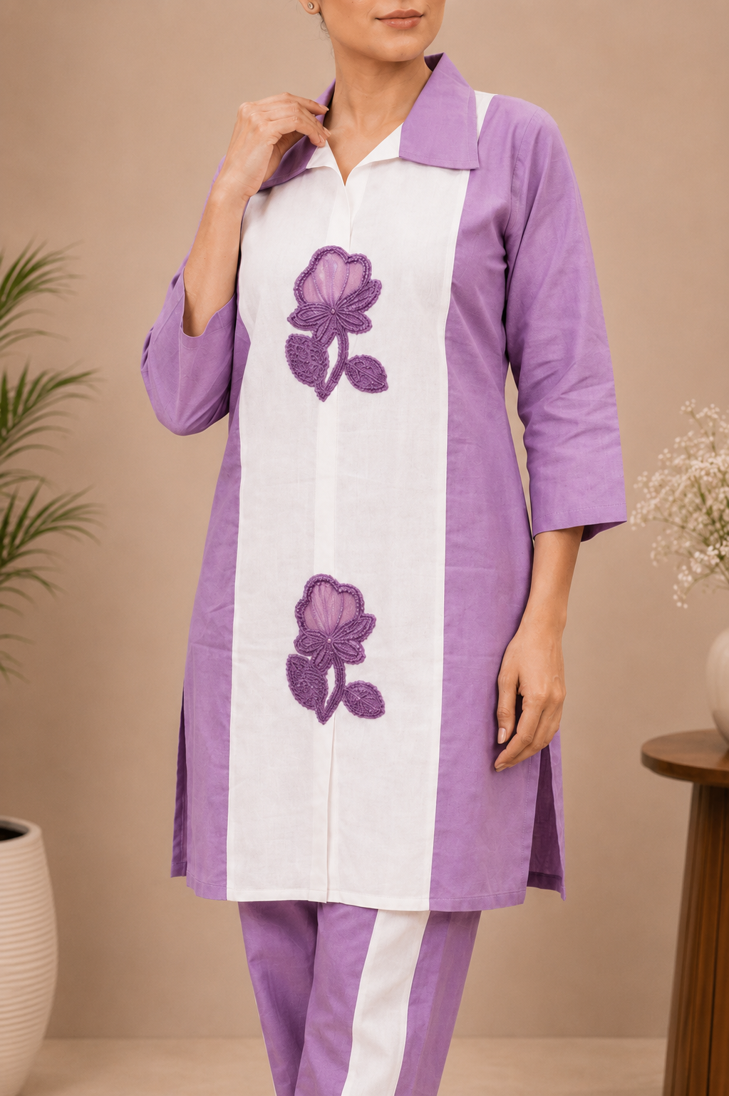 A woman modeling a lavender and white two-piece kurta set with purple floral embroidery, standing in a studio with beige walls and decorative plants.