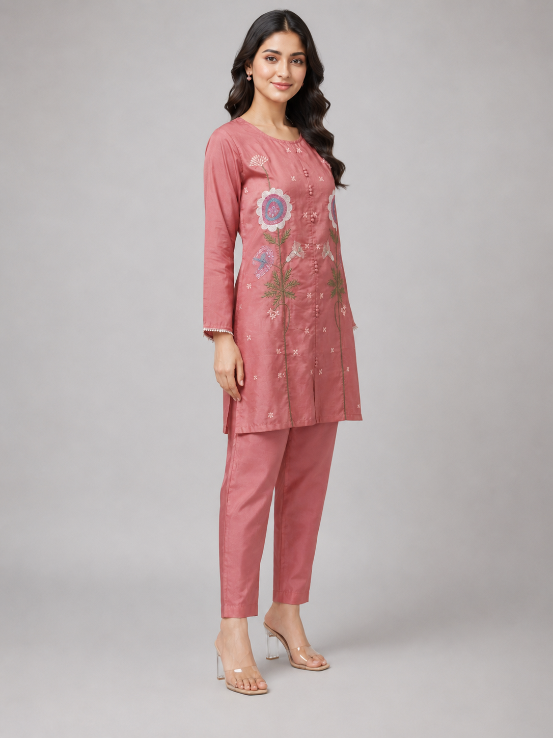 A woman modeling a dusty pink kurta and trouser set with floral embroidery on the front, photographed against a neutral gray studio background.
