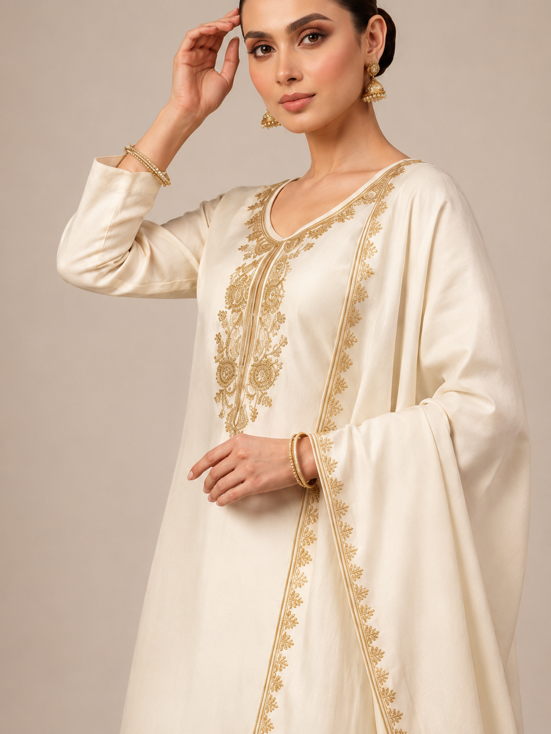 A woman modeling a traditional cream-colored Indian outfit consisting of a long kurta with gold embroidery, matching trousers, and a draped dupatta, set against a plain beige studio background.