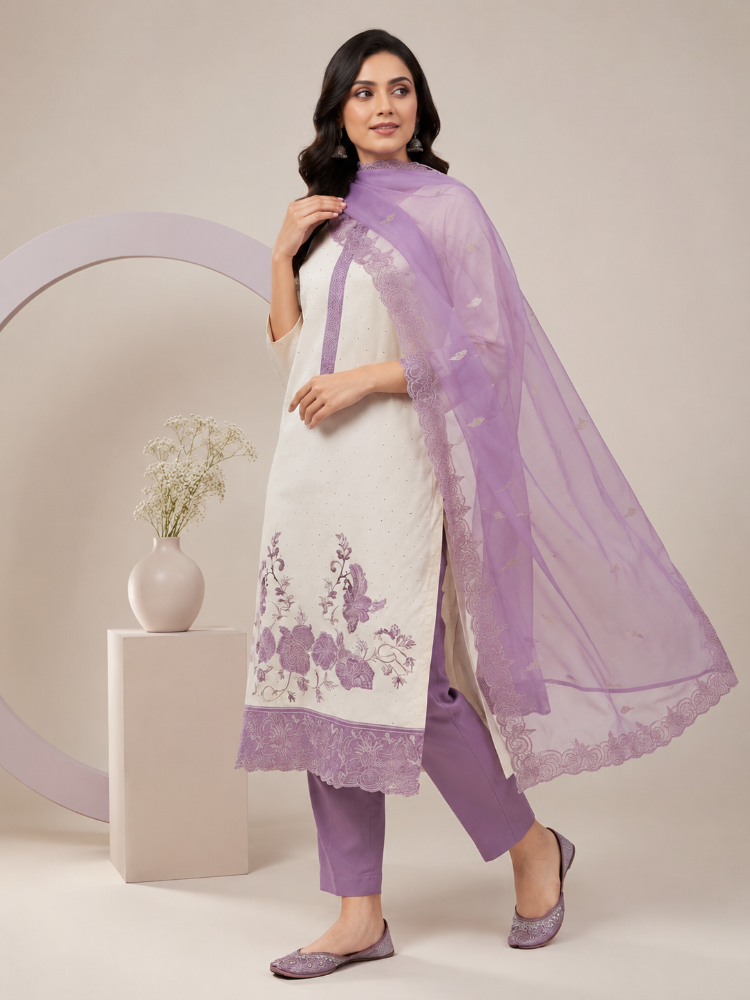 A woman models a traditional Indian salwar kameez suit set. The outfit features an off-white kurta with purple floral embroidery near the hem, paired with matching purple trousers and a sheer lavender dupatta draped over her shoulder.