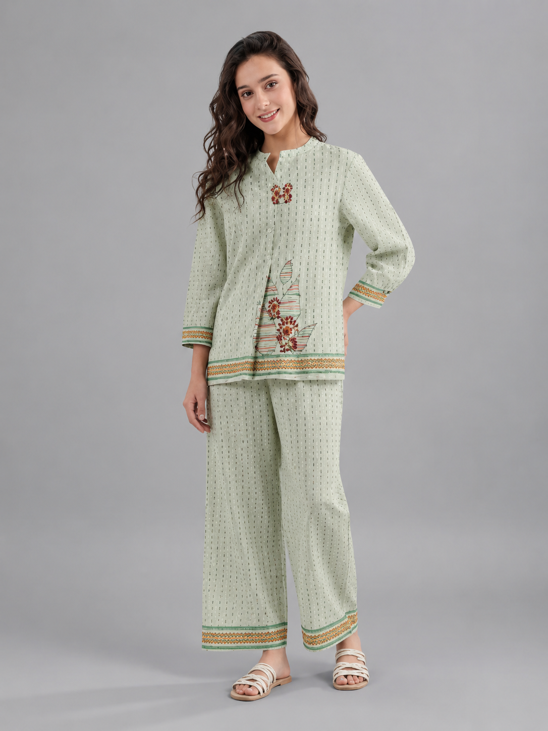 A woman modeling a light sage green two-piece kurta set with a small geometric pattern, featuring colorful embroidered details on the chest and sleeve cuffs, paired with matching wide-leg trousers.