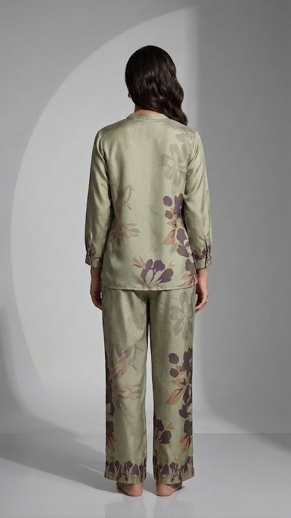 A woman modeling a two-piece satin pajama set in sage green with a floral print, posed against a gray studio background with a circular spotlight effect.