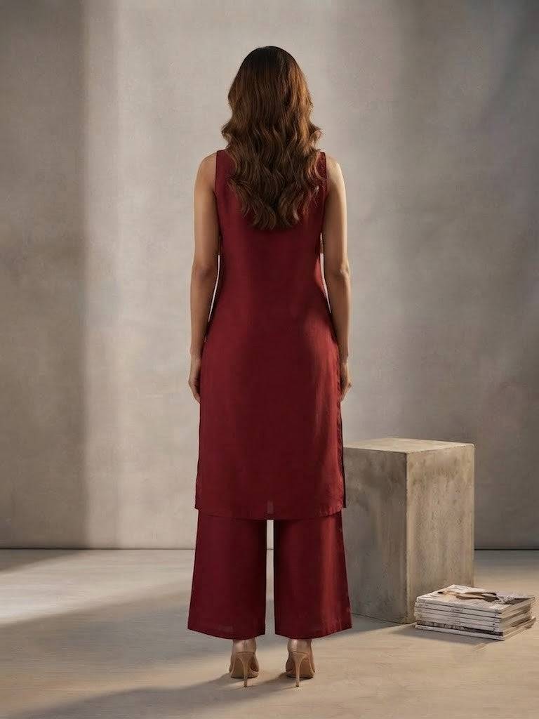 A woman models a deep red kurta set with wide-leg pants, featuring gold floral embroidery on the neckline, hem, and kurta front, set against a minimalist concrete block studio background.