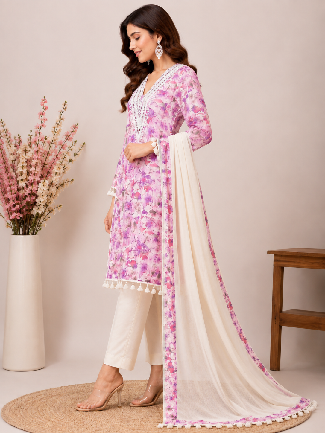 A woman models a traditional Indian salwar kameez suit set. The set features a light pink floral printed kurta with white tassel trim, white trousers, and a cream-colored dupatta with pink floral prints and a pink border.