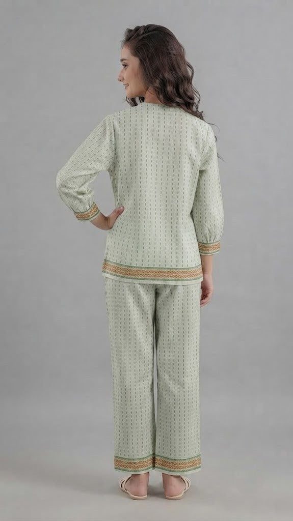 A woman modeling a light sage green two-piece kurta set with a small geometric pattern, featuring colorful embroidered details on the chest and sleeve cuffs, paired with matching wide-leg trousers.