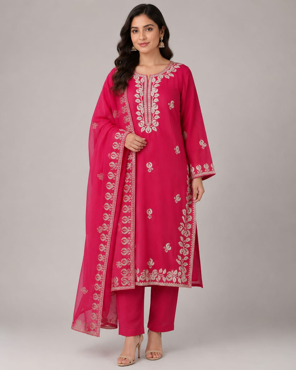 A woman modeling a bright pink traditional Indian outfit with a matching sheer dupatta, featuring intricate white floral embroidery on the kurta and dupatta, paired with matching pink trousers, against a neutral gray studio background.