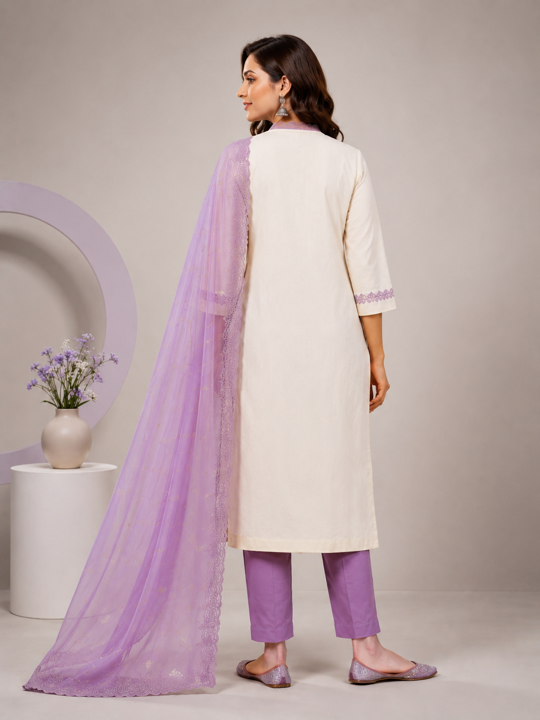 A woman models a traditional Indian salwar kameez suit set. The outfit features an off-white kurta with purple floral embroidery near the hem, paired with matching purple trousers and a sheer lavender dupatta draped over her shoulder.