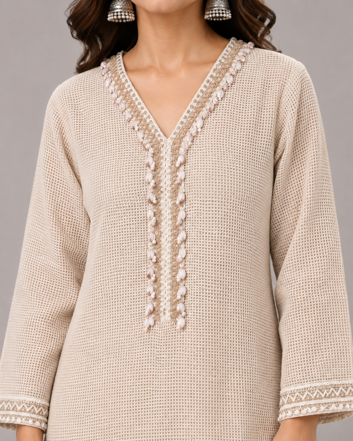 A woman modeling a light beige kurta set with matching trousers, featuring delicate gold embroidery on the neckline, cuffs, and hem, set against a solid warm beige studio background.