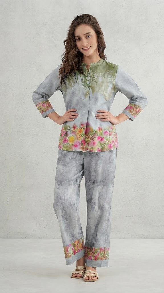 A woman modeling a two-piece traditional outfit featuring a light gray and white tie-dye kurta with green embroidery and colorful floral prints, paired with matching wide-leg trousers.