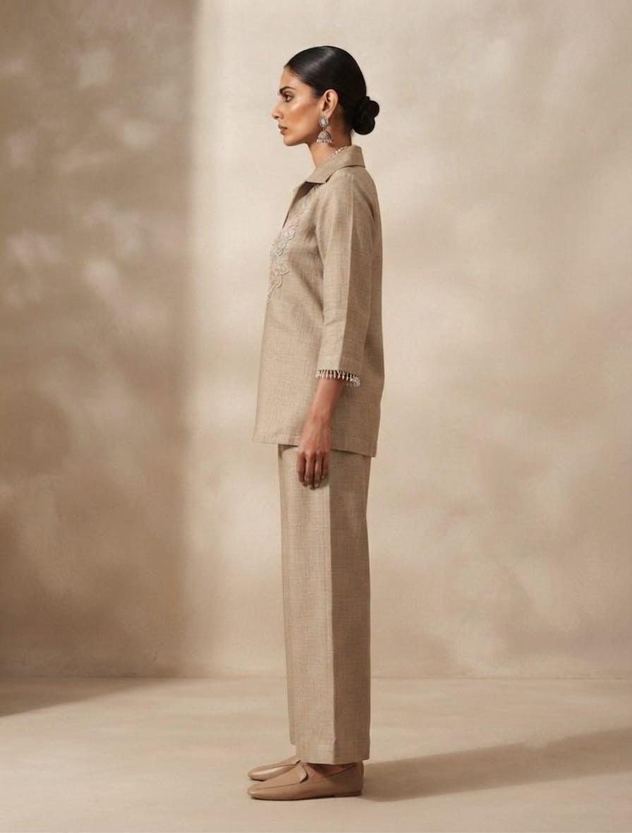 A woman stands against a warm beige textured backdrop wearing a matching beige two-piece outfit with a collared embroidered kurta and wide-leg trousers. The kurta has delicate floral embroidery around the neckline and three-quarter sleeves with ruffled cuffs.