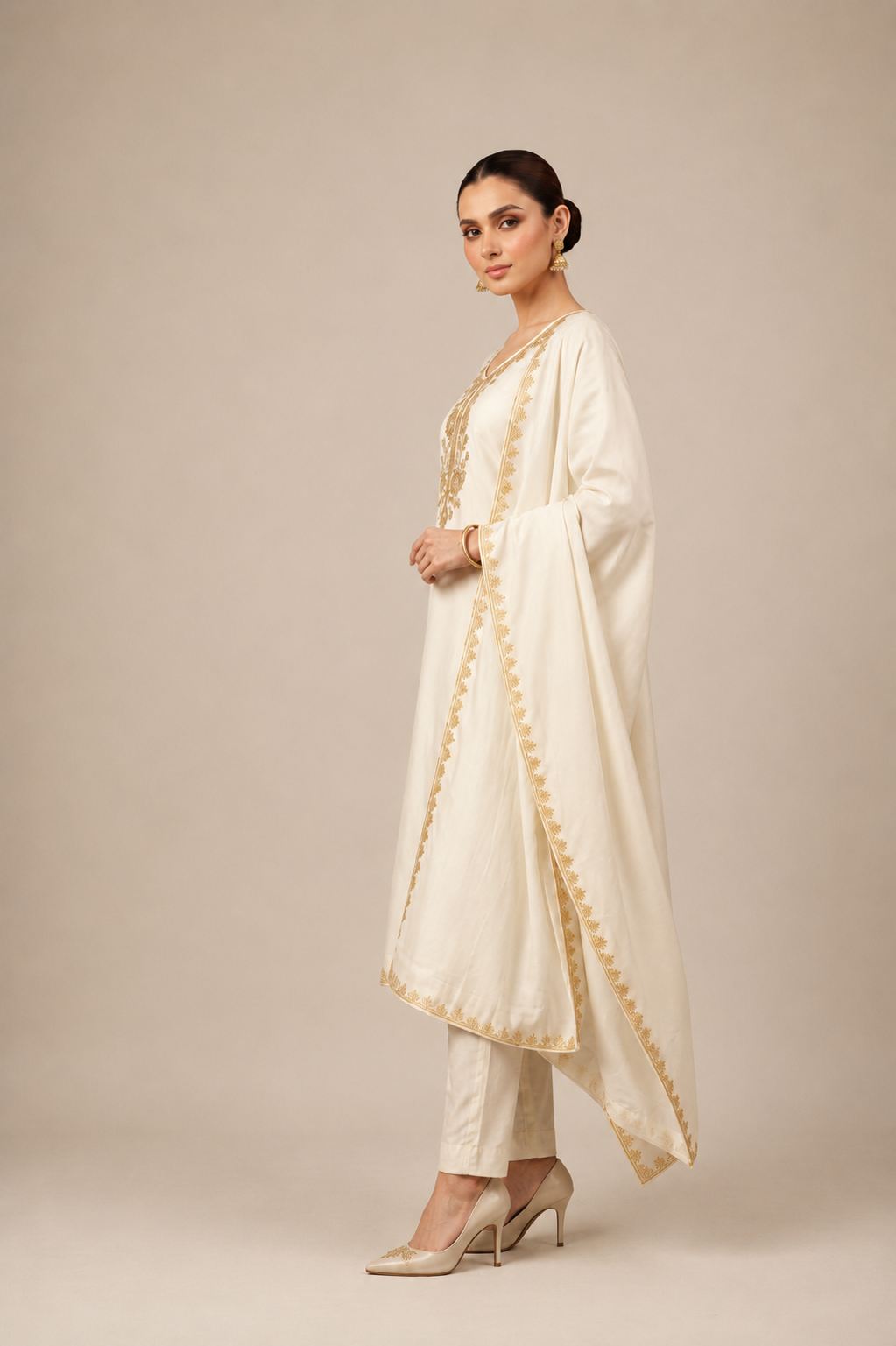 A woman modeling a traditional cream-colored Indian outfit consisting of a long kurta with gold embroidery, matching trousers, and a draped dupatta, set against a plain beige studio background.