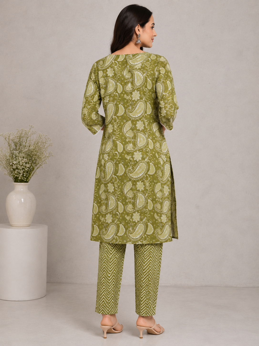 A woman models a green printed kurta set with matching patterned pants, standing in a studio setting with a neutral gray background and a small plant on a white pedestal.