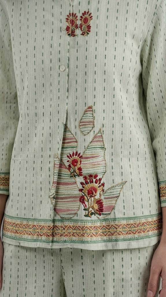 A woman modeling a light sage green two-piece kurta set with a small geometric pattern, featuring colorful embroidered details on the chest and sleeve cuffs, paired with matching wide-leg trousers.