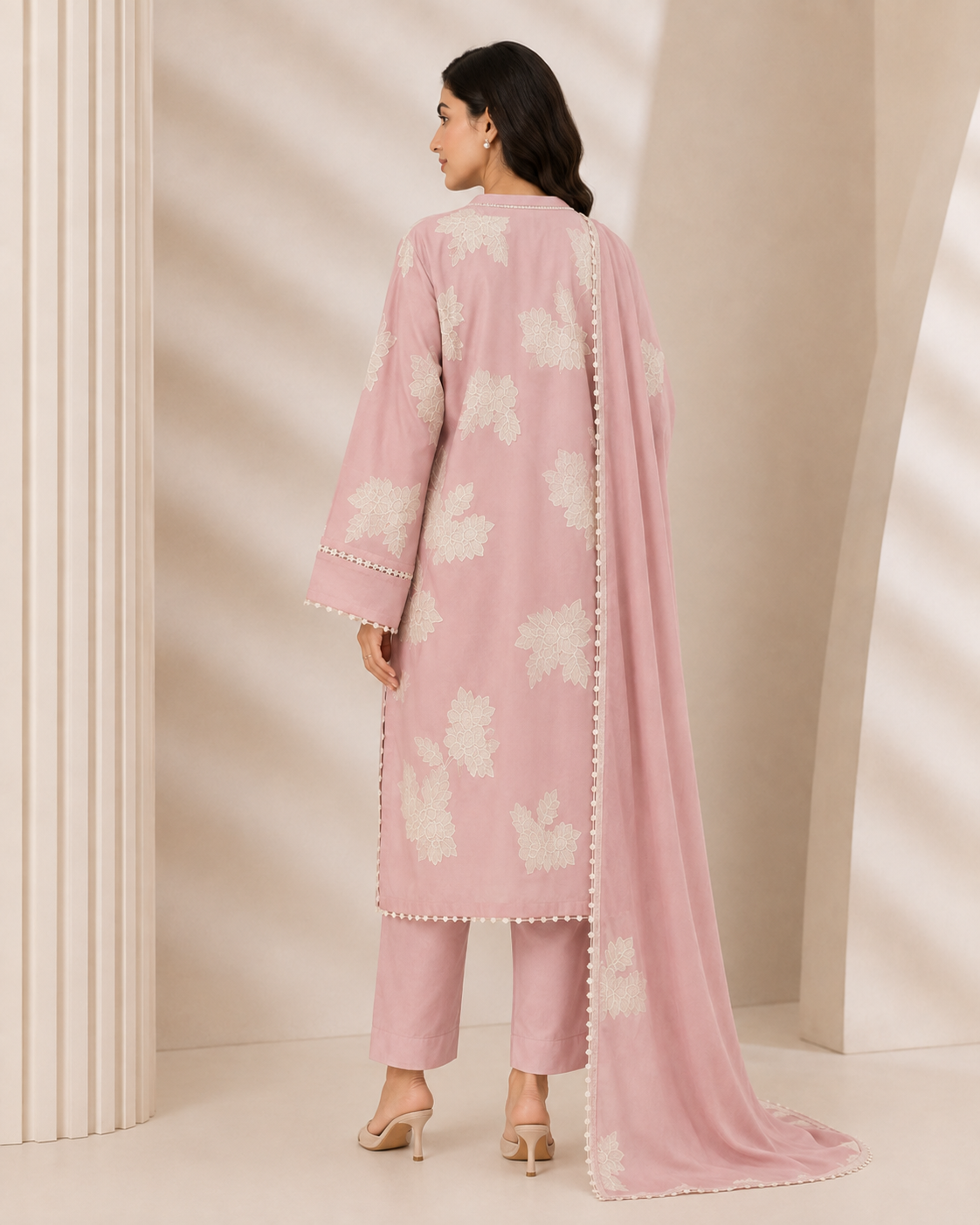 A woman models a three-piece traditional Indian outfit consisting of a dusty pink kurta with white floral embroidery, matching pink trousers, and a coordinating pink dupatta with white floral prints and pom-pom trim, set against a beige architectural background with soft shadows.