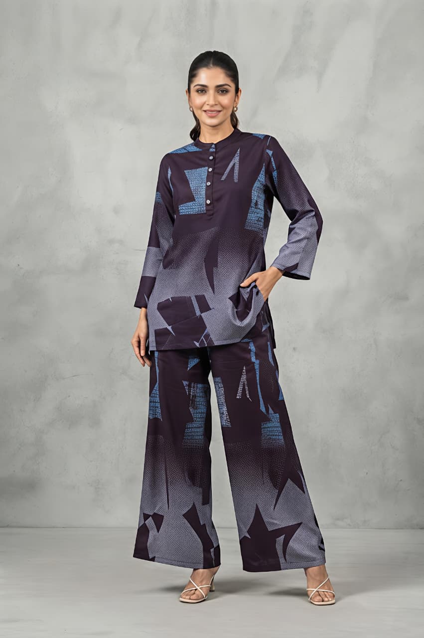 A woman modeling a matching two-piece printed outfit consisting of a long-sleeve button-up kurta and wide-leg trousers, featuring an abstract geometric pattern in shades of purple, gray, and blue, set against a textured gray studio backdrop.