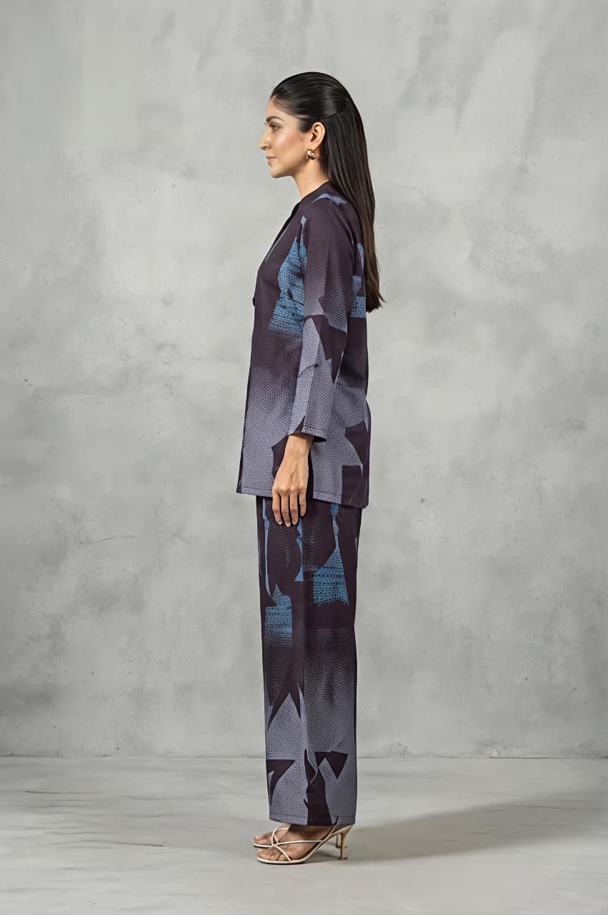 A woman modeling a matching two-piece printed outfit consisting of a long-sleeve button-up kurta and wide-leg trousers, featuring an abstract geometric pattern in shades of purple, gray, and blue, set against a textured gray studio backdrop.