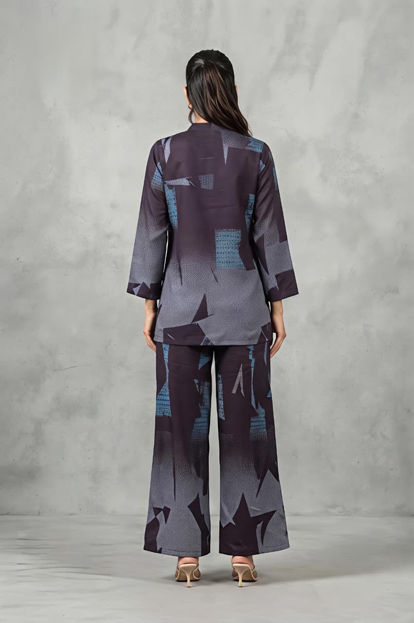 A woman modeling a matching two-piece printed outfit consisting of a long-sleeve button-up kurta and wide-leg trousers, featuring an abstract geometric pattern in shades of purple, gray, and blue, set against a textured gray studio backdrop.