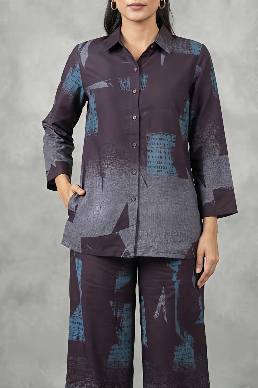 A woman modeling a matching two-piece printed outfit consisting of a long-sleeve button-up kurta and wide-leg trousers, featuring an abstract geometric pattern in shades of purple, gray, and blue, set against a textured gray studio backdrop.