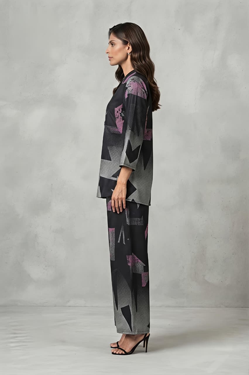 A woman models a matching two-piece set featuring a long-sleeve collared top and wide-leg trousers, both decorated with a modern geometric pattern in shades of gray, black, and pink, set against a textured gray studio backdrop.