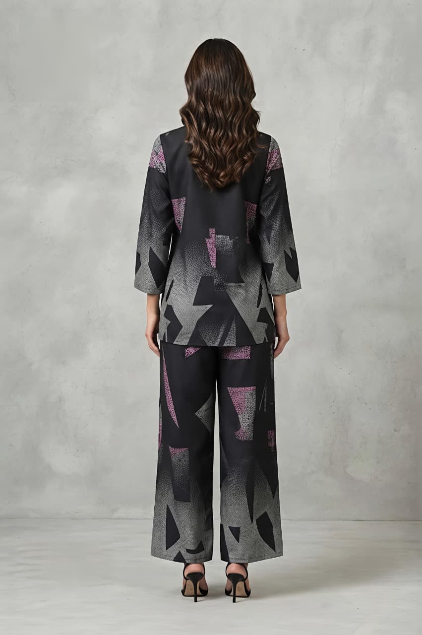 A woman models a matching two-piece set featuring a long-sleeve collared top and wide-leg trousers, both decorated with a modern geometric pattern in shades of gray, black, and pink, set against a textured gray studio backdrop.