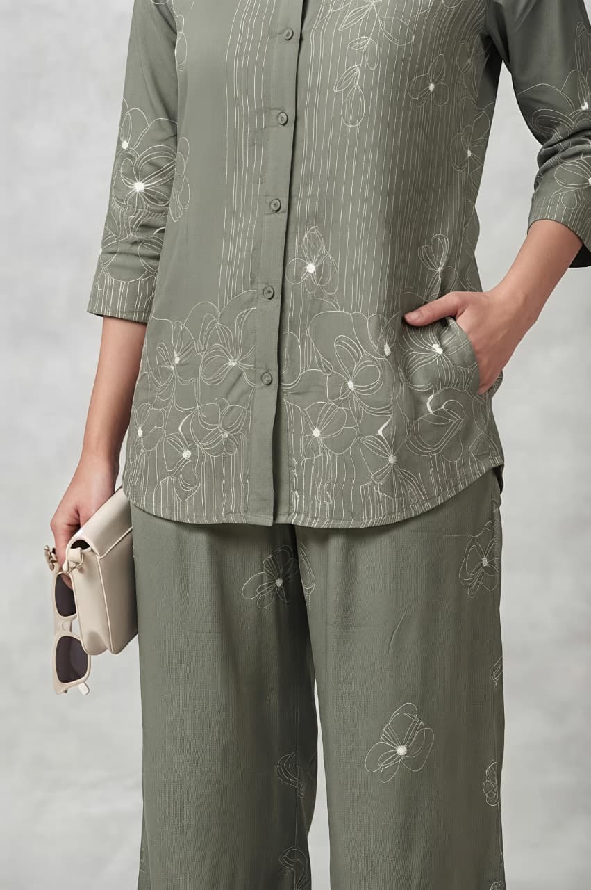 A woman models a matching two-piece outfit consisting of long-sleeve button-up shirt and wide-leg trousers, both decorated with white floral embroidery. She is holding a small beige clutch and wearing beige sandals against a neutral gray studio background.