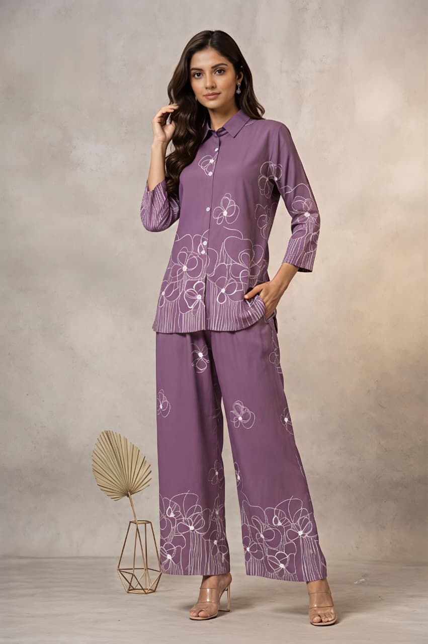 A woman models a matching purple two-piece kurta set with white floral embroidery, consisting of a long-sleeve button-up shirt and wide-leg trousers, set against a textured beige studio backdrop.