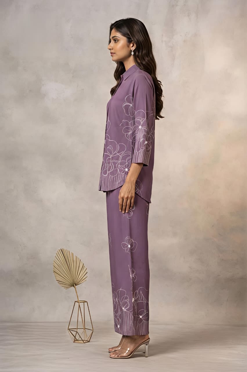 A woman models a matching purple two-piece kurta set with white floral embroidery, consisting of a long-sleeve button-up shirt and wide-leg trousers, set against a textured beige studio backdrop.