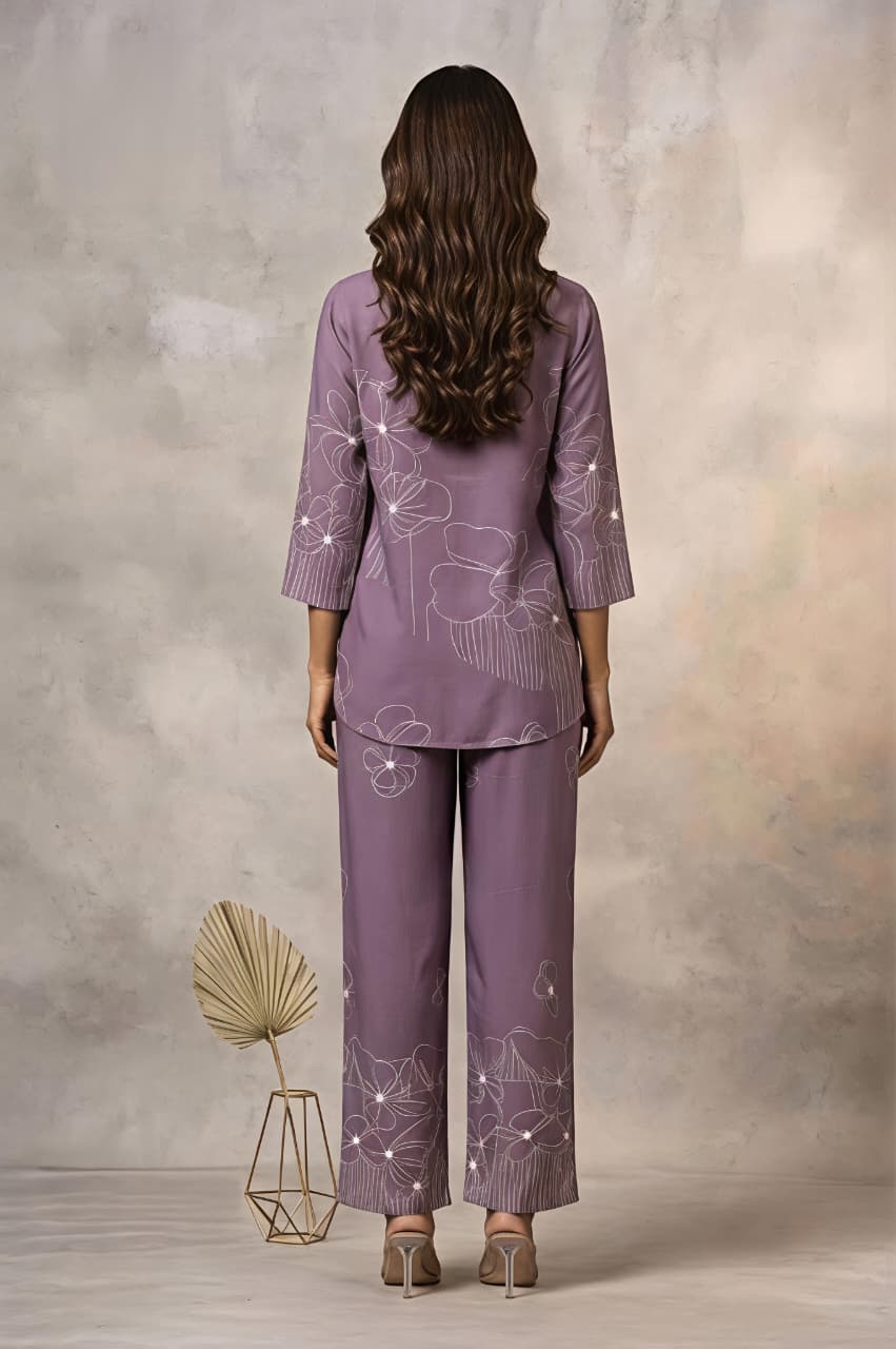 A woman models a matching purple two-piece kurta set with white floral embroidery, consisting of a long-sleeve button-up shirt and wide-leg trousers, set against a textured beige studio backdrop.
