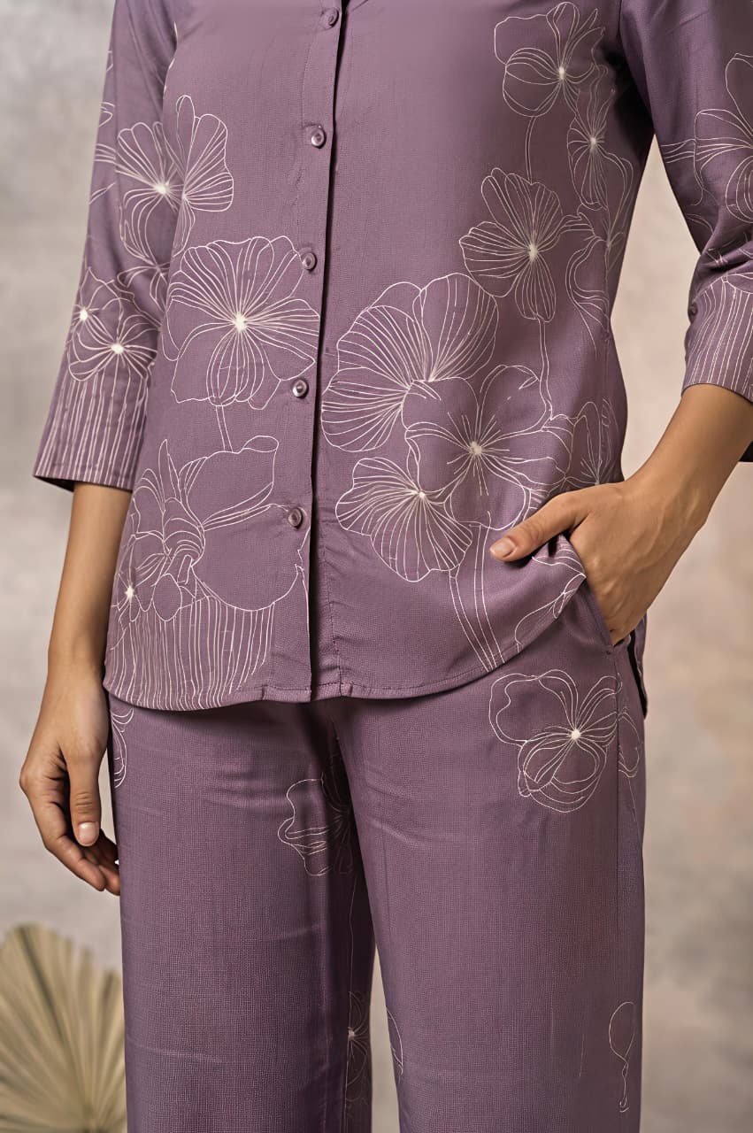 A woman models a matching purple two-piece kurta set with white floral embroidery, consisting of a long-sleeve button-up shirt and wide-leg trousers, set against a textured beige studio backdrop.