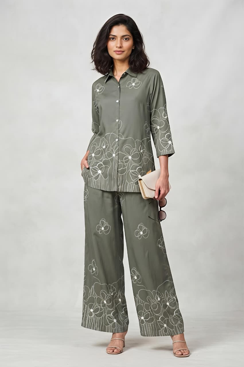 A woman models a matching two-piece outfit consisting of long-sleeve button-up shirt and wide-leg trousers, both decorated with white floral embroidery. She is holding a small beige clutch and wearing beige sandals against a neutral gray studio background.