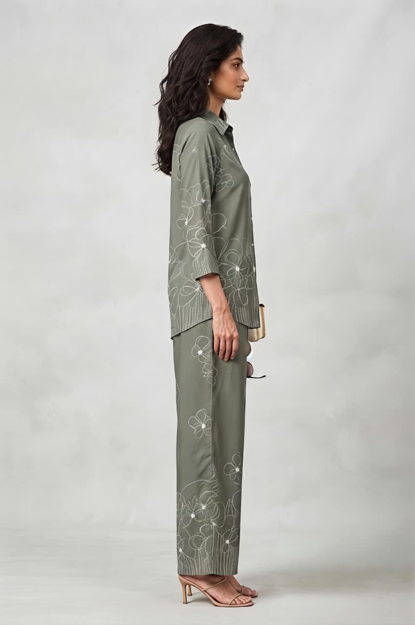 A woman models a matching two-piece outfit consisting of long-sleeve button-up shirt and wide-leg trousers, both decorated with white floral embroidery. She is holding a small beige clutch and wearing beige sandals against a neutral gray studio background.