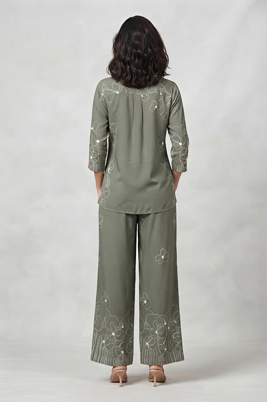 A woman models a matching two-piece outfit consisting of long-sleeve button-up shirt and wide-leg trousers, both decorated with white floral embroidery. She is holding a small beige clutch and wearing beige sandals against a neutral gray studio background.
