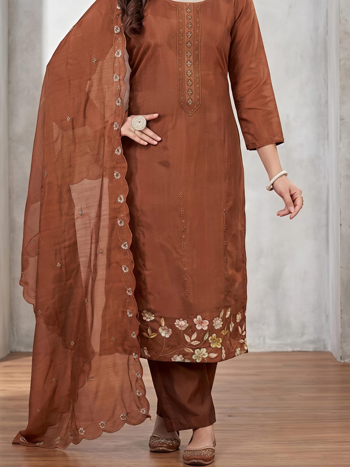 A woman models a brown traditional Indian salwar kameez suit set, featuring a brown kurta with floral embroidery at the hem, matching brown trousers, and a sheer brown dupatta with decorative buttons down the side, set against a gray concrete wall background.