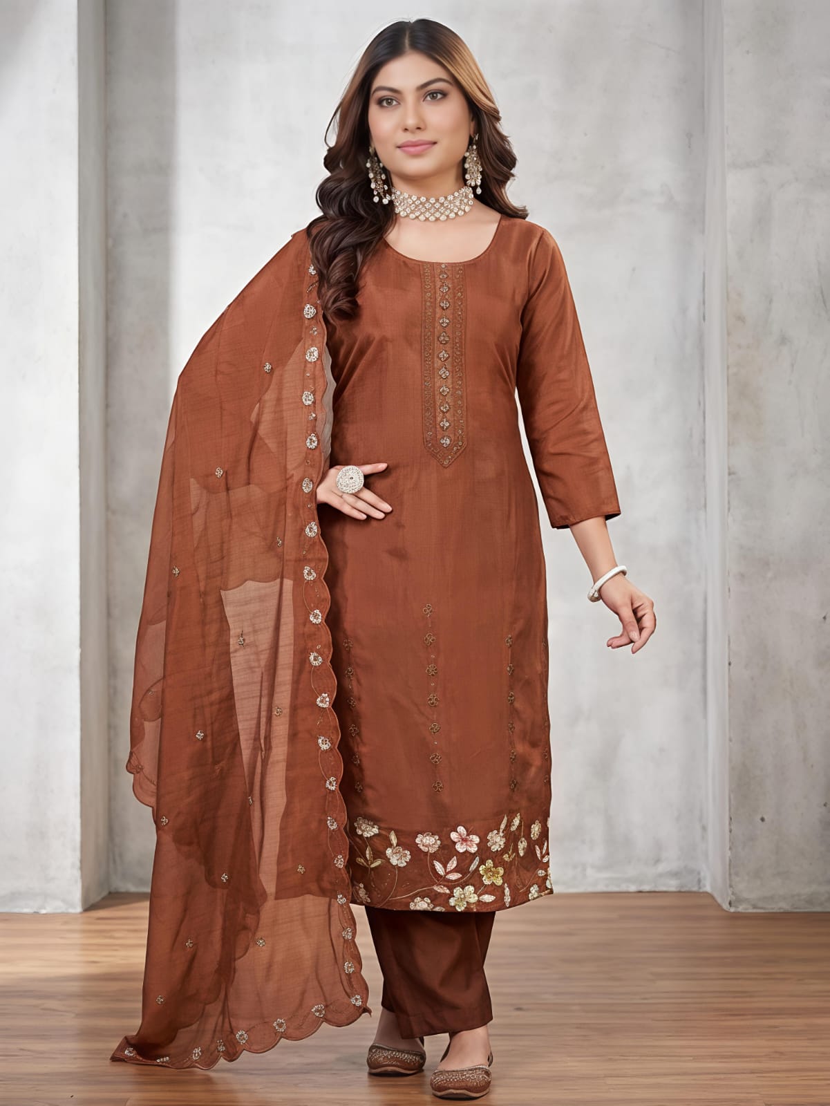A woman models a brown traditional Indian salwar kameez suit set, featuring a brown kurta with floral embroidery at the hem, matching brown trousers, and a sheer brown dupatta with decorative buttons down the side, set against a gray concrete wall background.