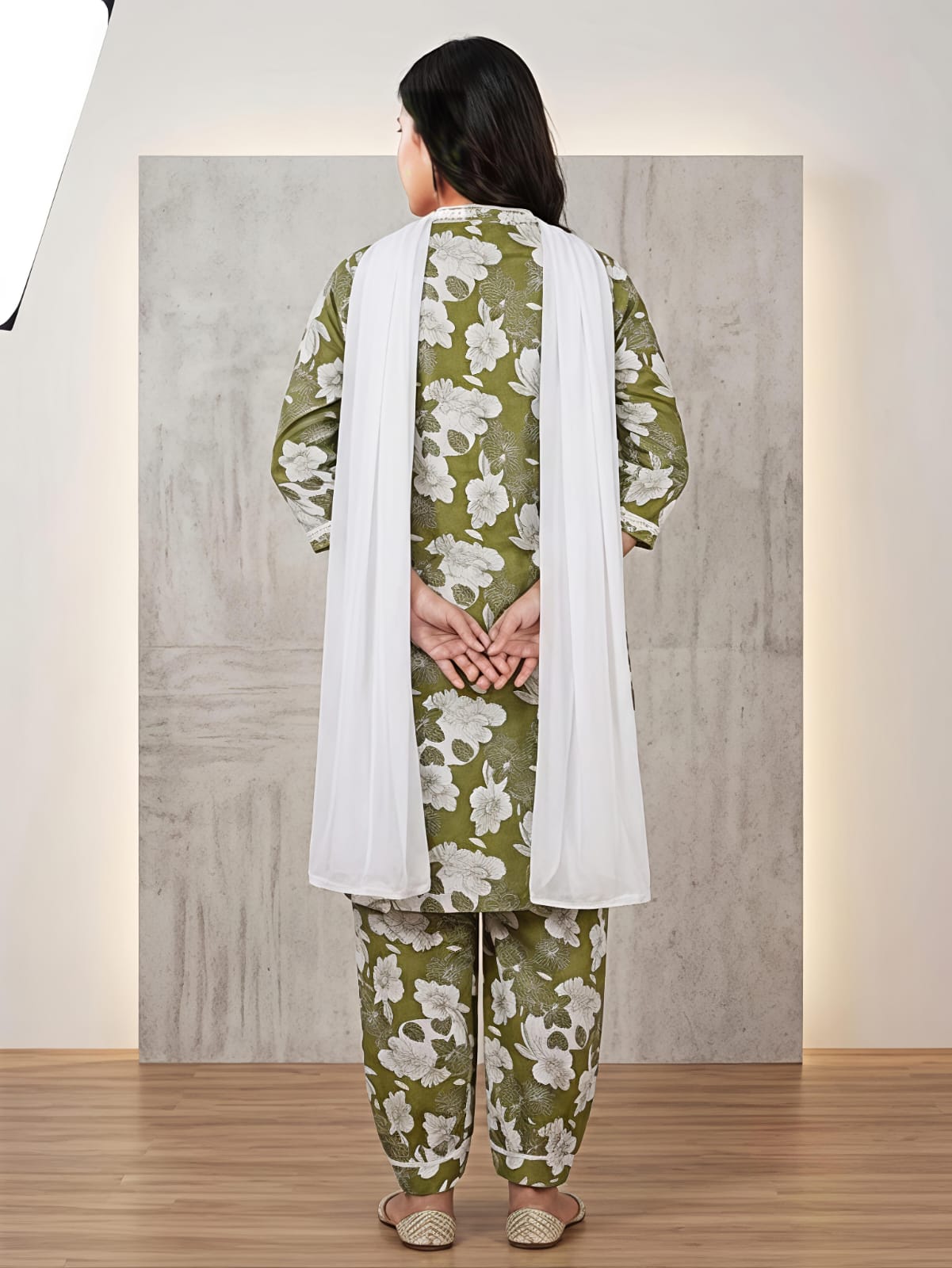 A woman modeling a green and white floral printed kurta set with matching pants and a white dupatta, standing in front of a textured gray backdrop.