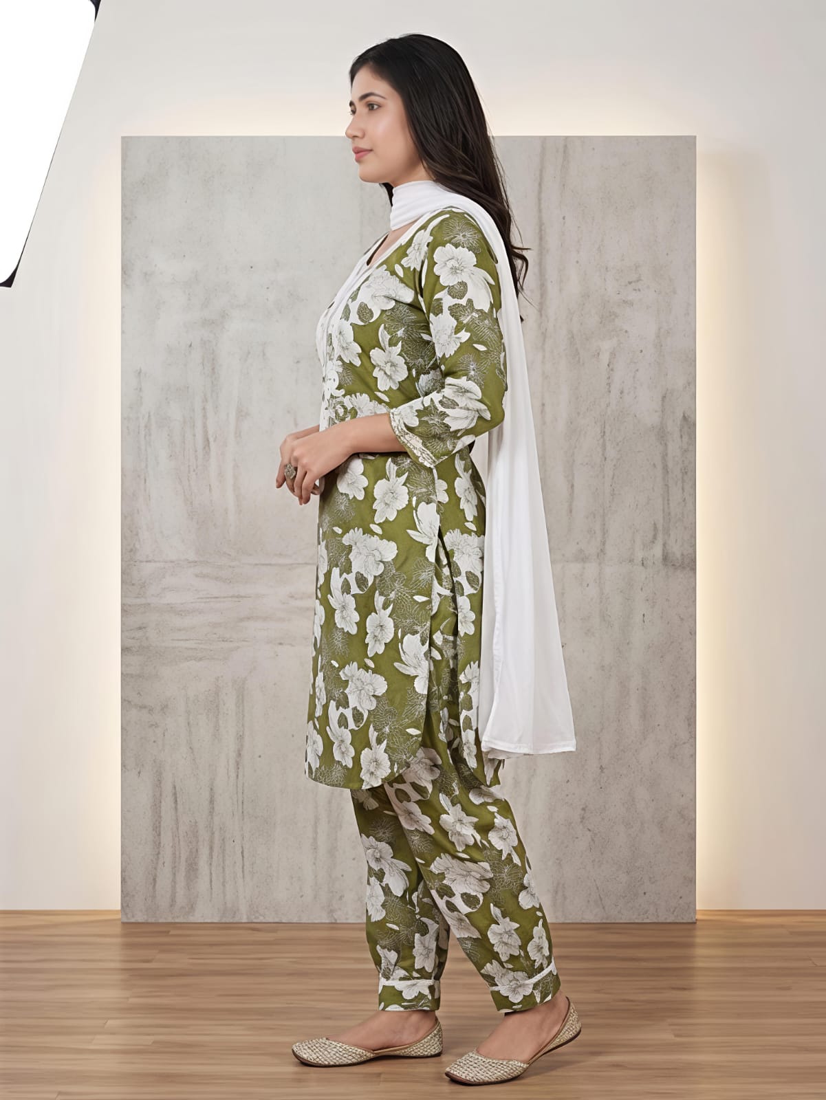 A woman modeling a green and white floral printed kurta set with matching pants and a white dupatta, standing in front of a textured gray backdrop.