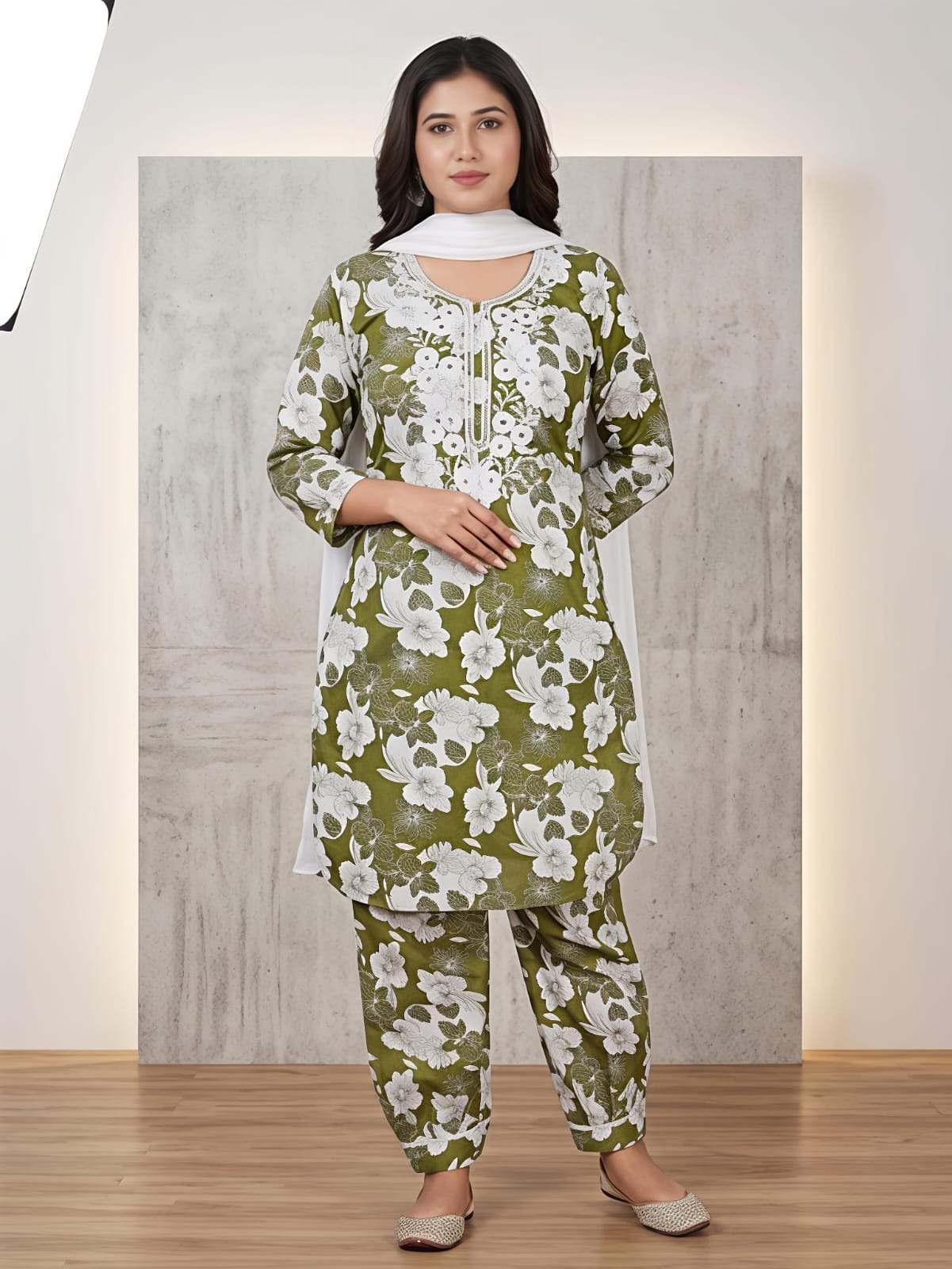 A woman modeling a green and white floral printed kurta set with matching pants and a white dupatta, standing in front of a textured gray backdrop.