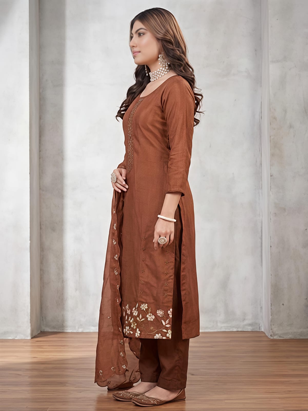 A woman models a brown traditional Indian salwar kameez suit set, featuring a brown kurta with floral embroidery at the hem, matching brown trousers, and a sheer brown dupatta with decorative buttons down the side, set against a gray concrete wall background.
